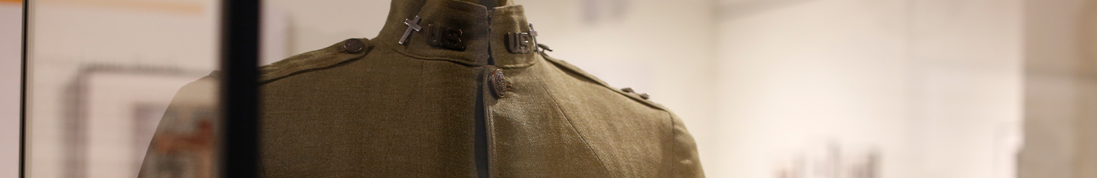 Cropped close-up focusing on the collar and shoulders of an olive-green military tunic with a cross insignia pin and other rank markers.