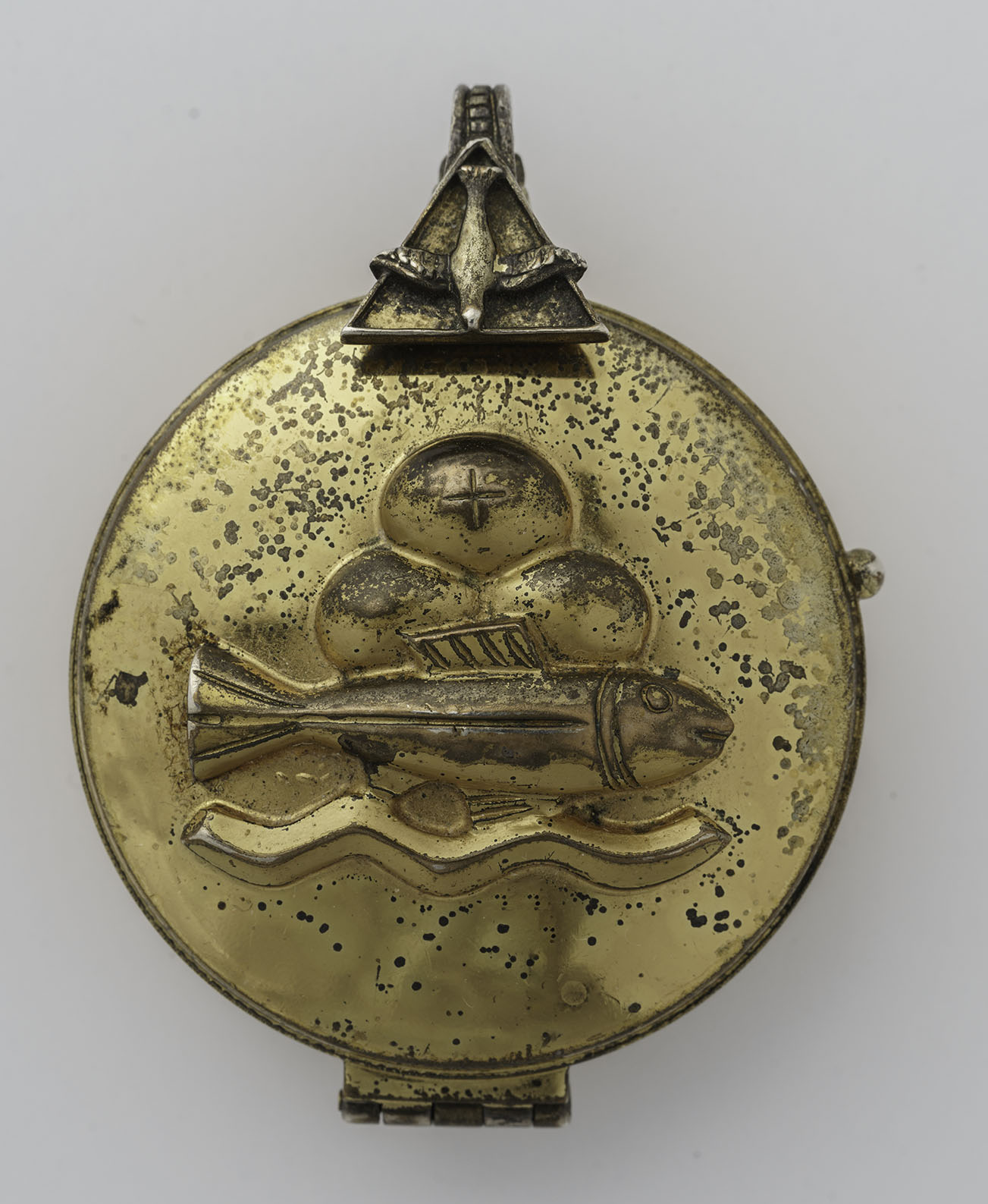 Modern photograph of a round golden container with a hinged lid and clasp. The clasp is decorated with the image of the Holy Spirit dove in a triangle and the lid depicts three loaves of bread (one with a cross), a fish, and water.